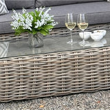 Hill Interiors Outdoor Large Corner Set in beige with woven rattan base, glass top holding two white wine glasses, small bowls and a vase of white flowers.