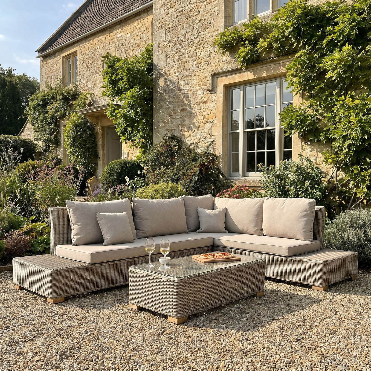 Hill Interiors Large Outdoor Corner Sofa and Coffee Table Set in beige woven rattan with glass-topped table, arranged on gravel outside a stone cottage.