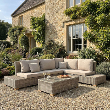 Hill Interiors Large Outdoor Corner Sofa and Coffee Table Set in beige woven rattan with glass-topped table, arranged on gravel outside a stone cottage.