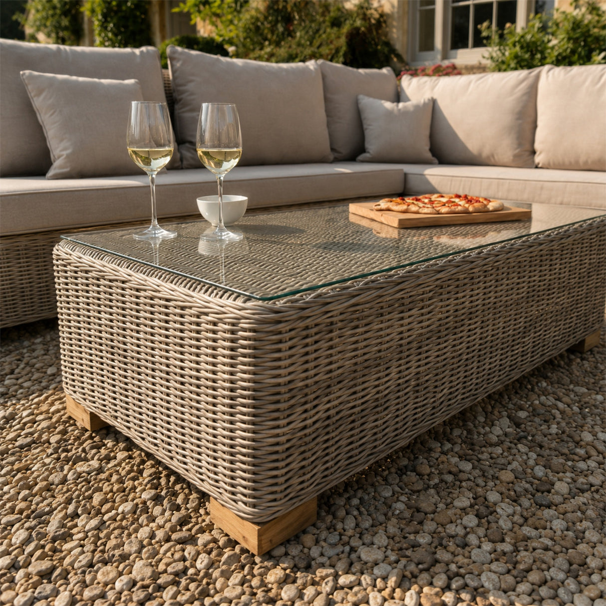 Hill Interiors beige outdoor corner sofa with glass-topped woven coffee table holding two white wine glasses and a pizza board on a gravel patio.