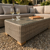 Hill Interiors beige outdoor corner sofa with glass-topped woven coffee table holding two white wine glasses and a pizza board on a gravel patio.