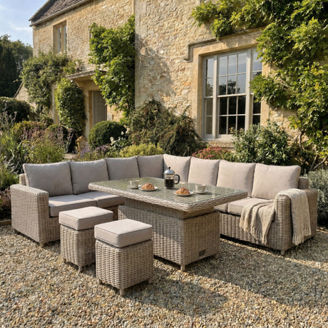 Hill Interiors Ferranock beige rattan corner sofa set with glass-topped riser table and two stools on gravel by a stone cottage garden.