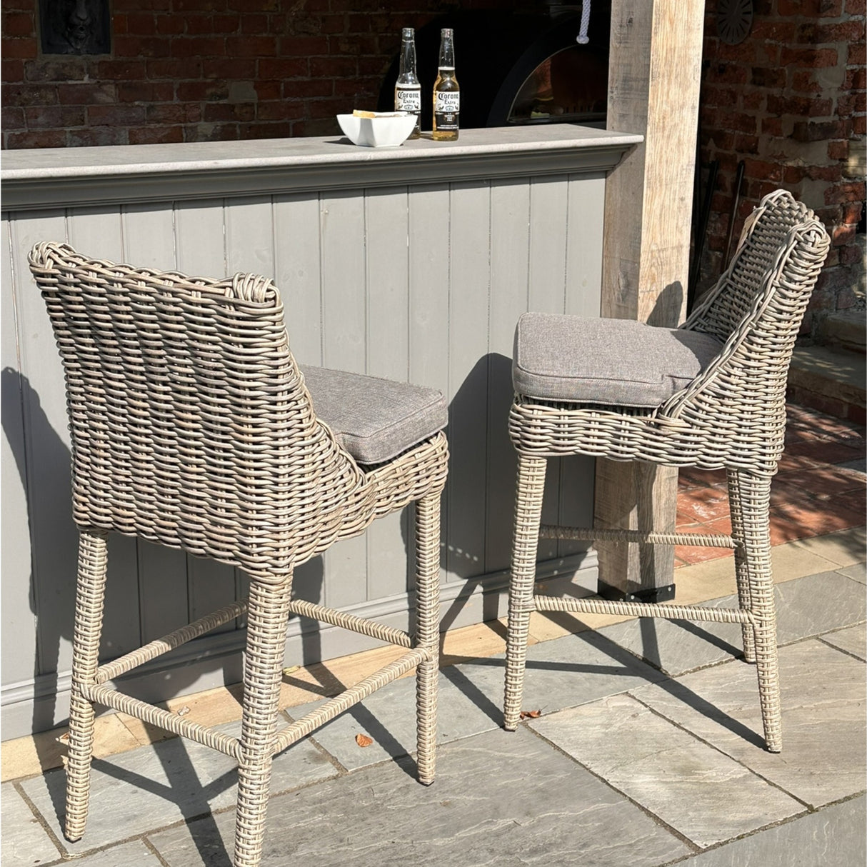Pair of Hill Interiors Metal Outdoor Bar Stools in beige with woven rattan-effect frames and grey cushions, set at a painted outdoor bar on a stone patio.