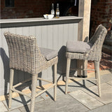 Pair of Hill Interiors Metal Outdoor Bar Stools in beige with woven rattan-effect frames and grey cushions, set at a painted outdoor bar on a stone patio.
