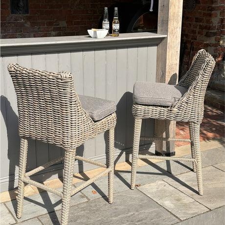 Pair of Hill Interiors Metal Outdoor Bar Stools in beige with woven rattan-effect frames and grey cushions, set at a painted outdoor bar on a stone patio.