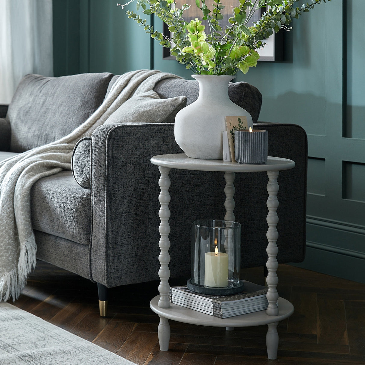 Hill Interiors Round Wooden Side Table in grey with turned spindle legs and lower shelf, styled beside a grey sofa with a vase and candle on top.