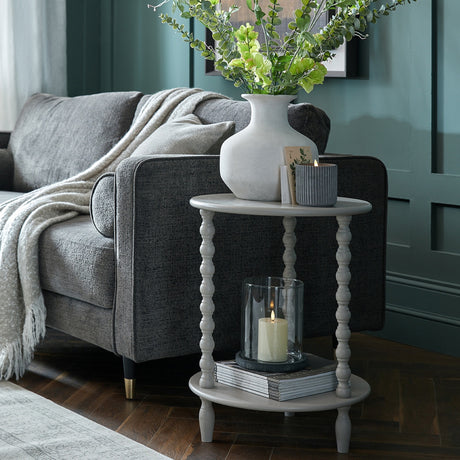Hill Interiors Round Wooden Side Table in grey with turned spindle legs and lower shelf, styled beside a grey sofa with a vase and candle on top.