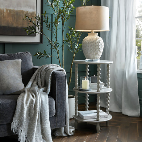 Hill Interiors Wooden Three-Tier Side Table in grey styled beside a dark sofa with a woven ceramic lamp, candles and books in a green living room.