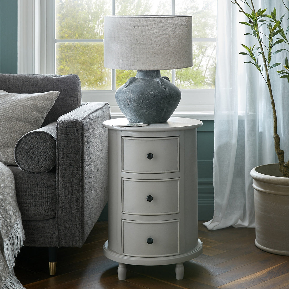 Hill Interiors 3 Drawer Round Side Table in grey wood beside a grey sofa, topped with a stone lamp in a bright living room by the window.