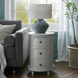 Hill Interiors 3 Drawer Round Side Table in grey wood beside a grey sofa, topped with a stone lamp in a bright living room by the window.