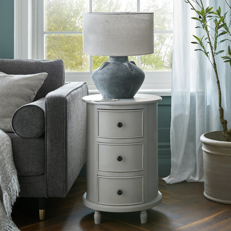 Hill Interiors 3 Drawer Round Side Table in grey wood beside a grey sofa, topped with a stone lamp in a bright living room by the window.