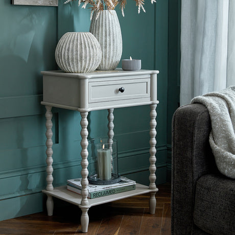 Hill Interiors Wooden Bobbin-Leg Side Table in grey with single drawer and lower shelf, styled with vases and candles beside a sofa in a green room.