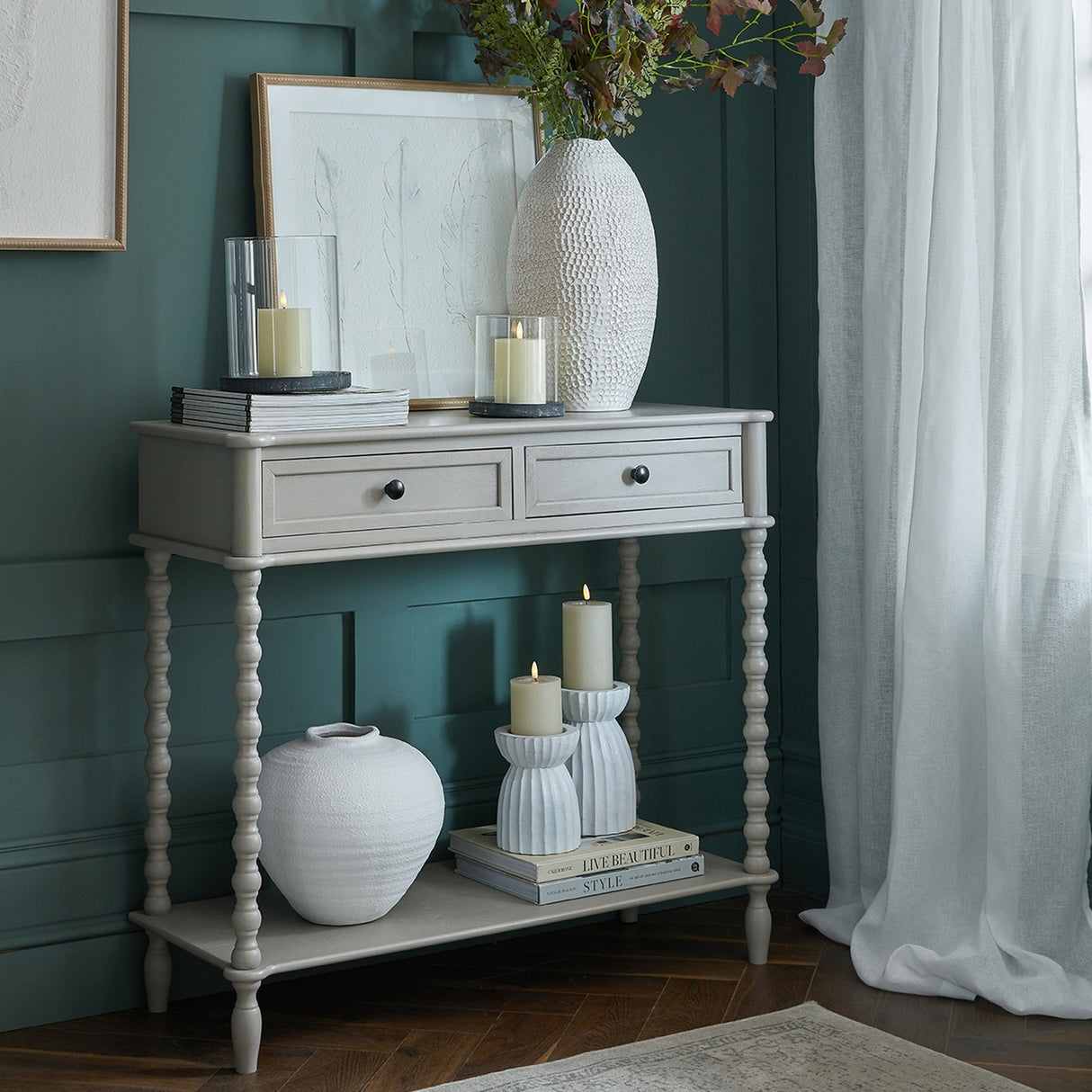 Hill Interiors Two Drawer Console Table in grey wood with turned legs and lower shelf, styled with vases and candles against a green panelled wall.