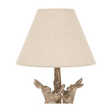 Hill Interiors Marching Hares table lamp in brown resin with sculpted hares at the base and a tapered natural linen shade, on a white background.