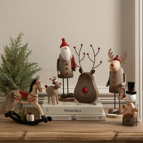 Hill Interiors Rustic Tall Metal Santa Figure in brown, styled on books alongside wooden reindeer, snowman, rocking horse and a small potted tree.
