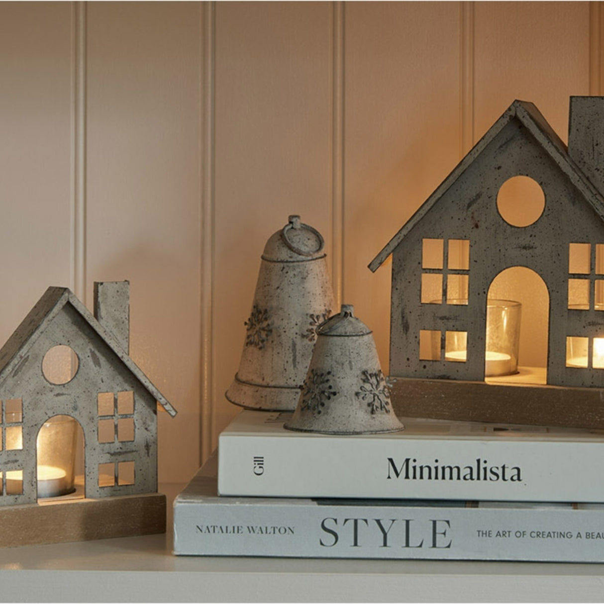Hill Interiors rustic white metal hanging bells with snowflake motifs, displayed on stacked style books beside wooden house tealight lanterns.