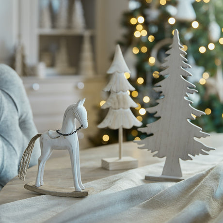 White-washed wooden rocking horse ornament by Hill Interiors displayed with carved white trees beside a lit Christmas tree on a linen-draped table.