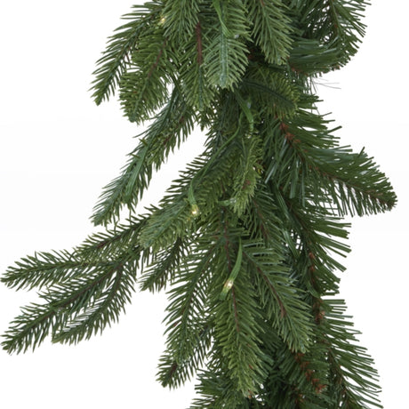 Close-up of Hill Interiors Pre-Lit Woodland Fir Christmas Garland in green, showing dense plastic needles and small warm LED lights on a white background.