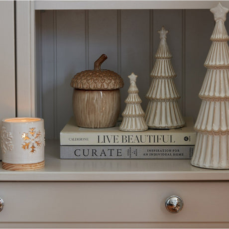 Hill Interiors medium ceramic snowy layered tree with star in white, styled on stacked books beside an acorn jar and taller ceramic trees.