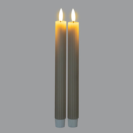 Pair of Hill Interiors Ribbed Taper Candles in beige wax with fine vertical ribbing and lit wicks, shown upright on a light grey background.