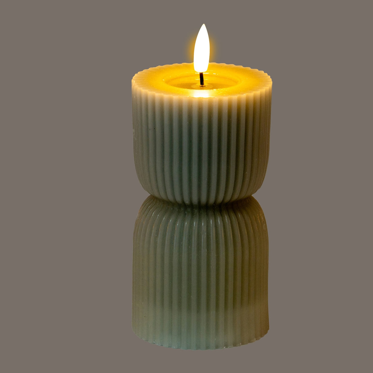 Hill Interiors Natural Glow carved LED pillar candle in beige with ribbed wax-effect finish and flickering flame, shown on a reflective grey surface.