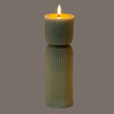 Hill Interiors Natural Glow carved LED pillar candle in beige, with ribbed plastic body and lit flame-effect tip, shown on a grey background.