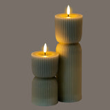 Hill Interiors Natural Glow carved LED pillar candles in beige, shown in two heights with ribbed texture and glowing flame-effect tips on a grey background.