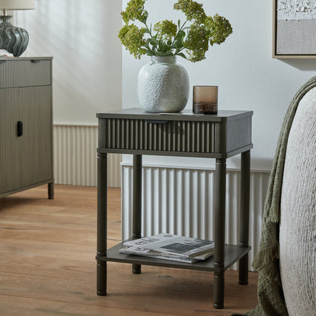 Hill Interiors 1 Drawer Wooden Side Table in grey with fluted drawer front and lower shelf, styled beside a bed with a vase of greenery and glass tumbler.