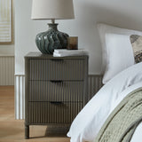 Hill Interiors 3 Drawer Wooden Bedside Table in brown with fluted drawer fronts, styled beside a bed with a teal ceramic lamp and stacked books on top.