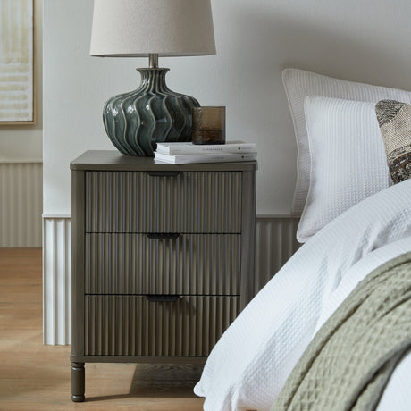 Hill Interiors 3 Drawer Wooden Bedside Table in brown with fluted drawer fronts, styled beside a bed with a teal ceramic lamp and stacked books on top.