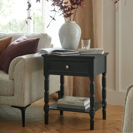 Hill Interiors Wooden One-Drawer Side Table in black with turned legs and lower shelf, styled beside a sofa with books, a glass and a ceramic vase.