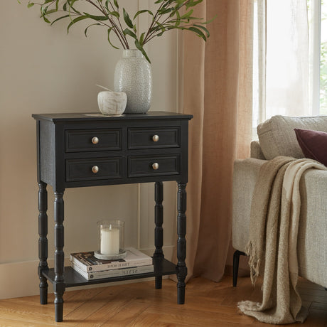 Hill Interiors 4 Drawer Side Table in black wood with turned legs and lower shelf, styled beside a sofa with vases, books and a candle.