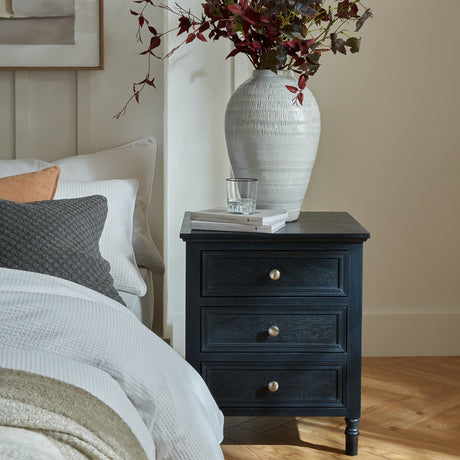 Hill Interiors Wooden Three-Drawer Side Table in black beside a bed, styled with books, a glass and a white ceramic vase of burgundy foliage.
