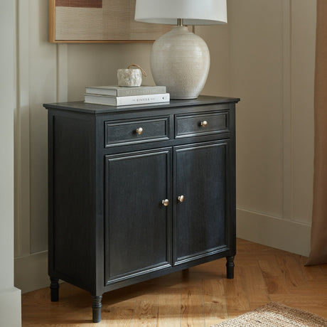 Hill Interiors black wooden two-door two-drawer sideboard styled in a hallway with a ceramic lamp, books and framed art above.