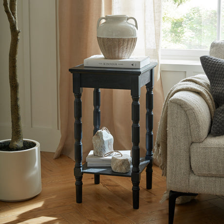 Hill Interiors Contour Wooden Side Table in black with turned legs and lower shelf, styled beside a cream sofa with books and a ceramic vase on top.