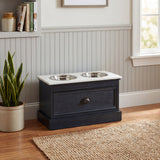 Hill Interiors wooden pet feeder table in black with white top and two stainless steel bowls, styled in a hallway with wainscoting and a snake plant.