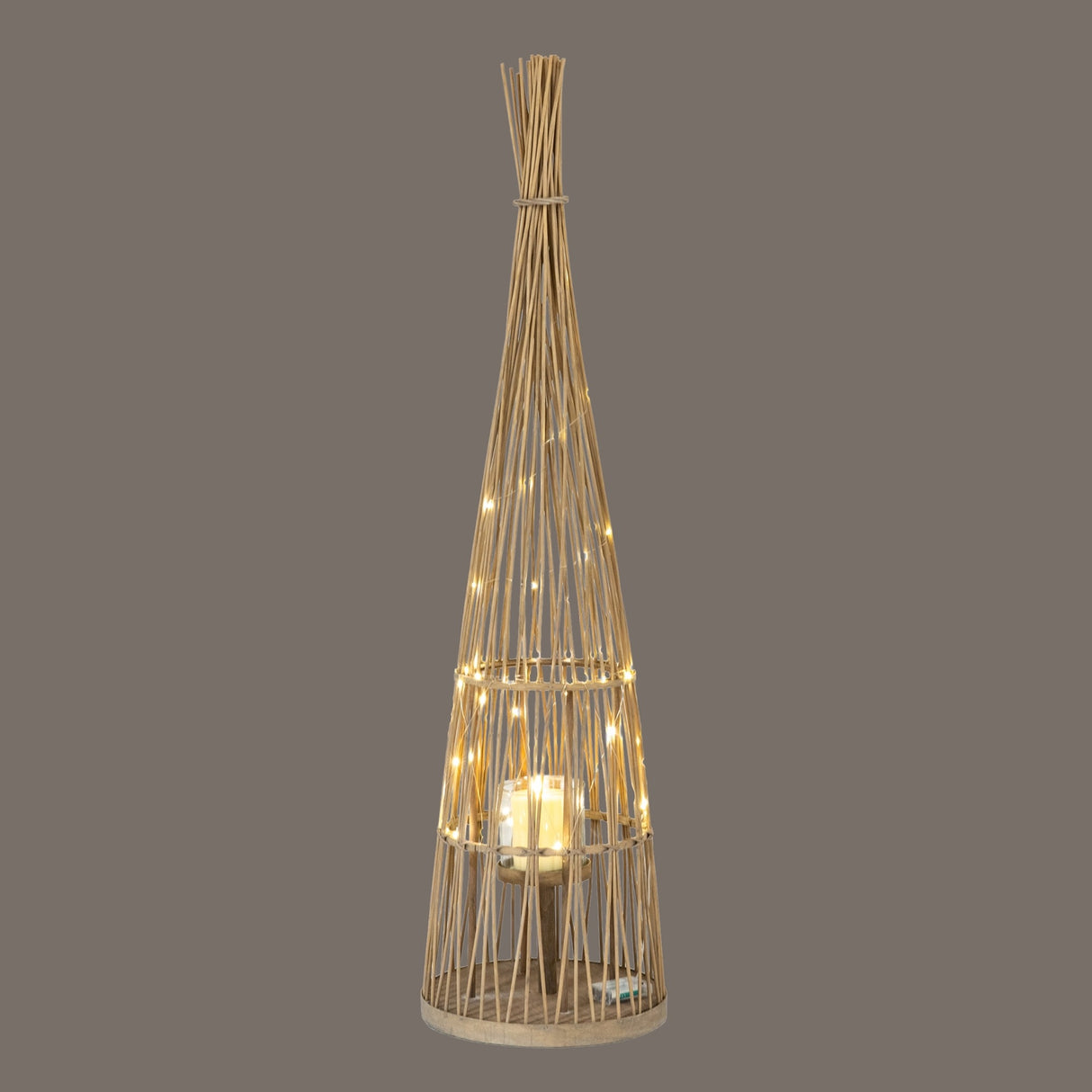 Hill Interiors large conical wicker lantern in brown with woven tapered frame, glass candle holder and lit LED fairy lights on a grey background.