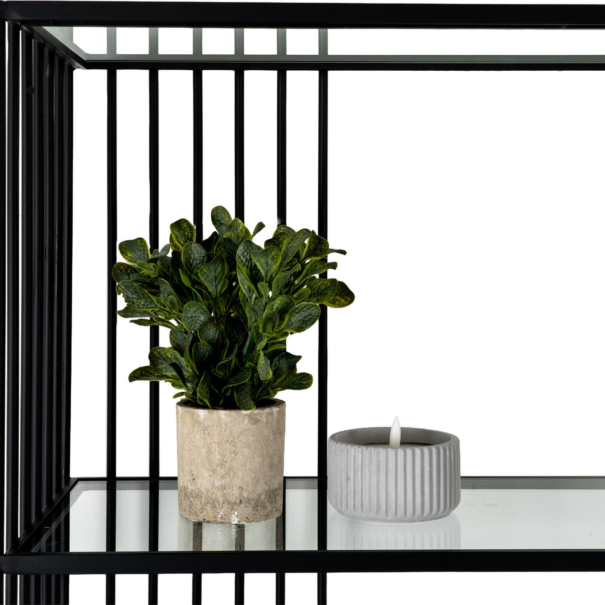 Hill Interiors Glass Shelf Unit with Black Metal Frame styled with a potted green plant and ribbed grey tealight holder on a clear tempered glass shelf.
