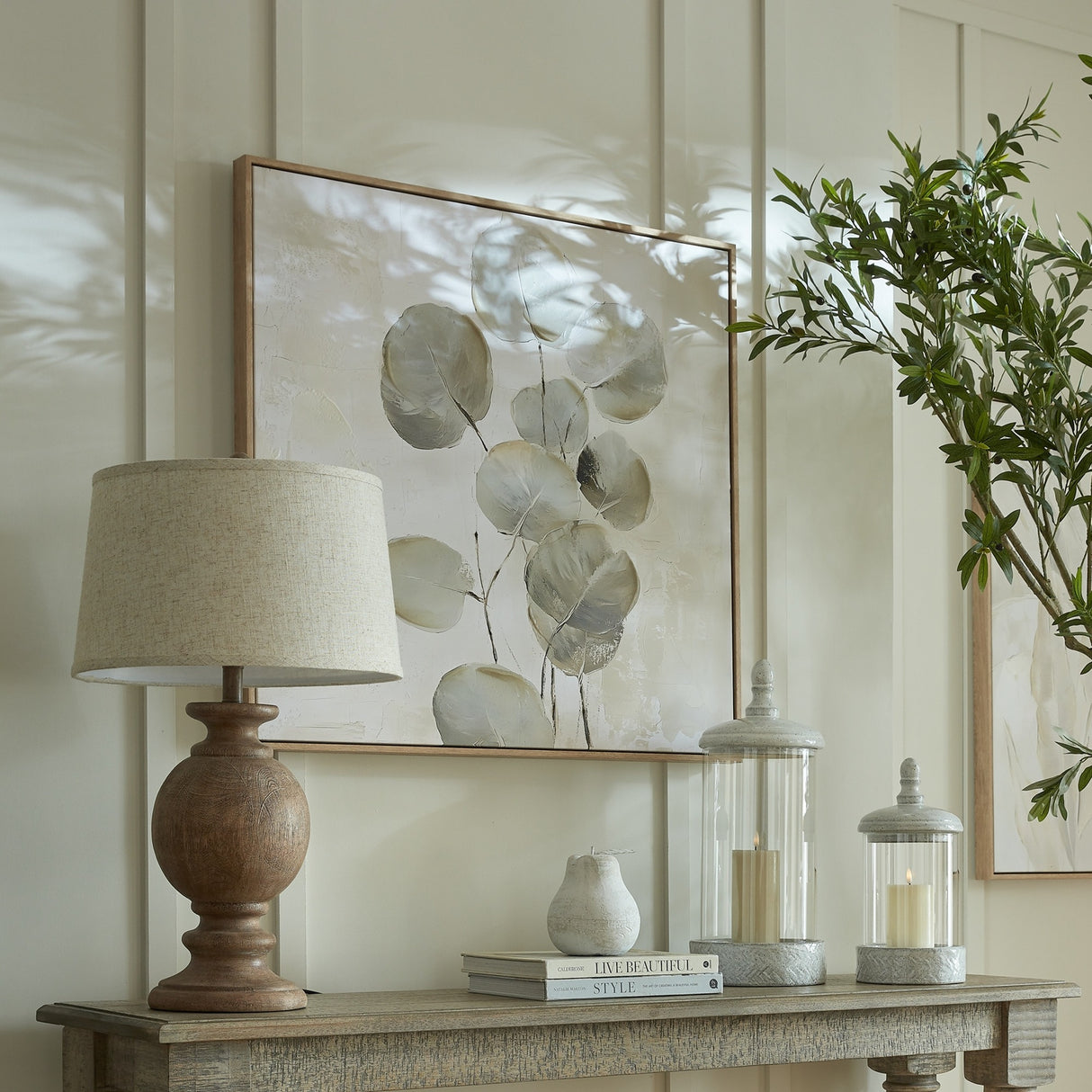 Hill Interiors table lamp with sculpted brown resin base and edged linen shade, styled on a console with botanical artwork, candle lanterns, and olive foliage.