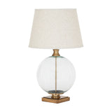 Hill Interiors Glass Orb Table Lamp with clear round base, brass-finished detailing and tapered natural linen shade with edged trim on white.