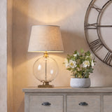 Hill Interiors Glass Orb Table Lamp with Edged Linen Shade in brass, lit on a wood sideboard beside white flowers and a metal wall clock.
