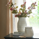 Hill Interiors Medium Hewn Ceramic Vase in white with textured body holding eucalyptus stems, set on stacked books beside a smaller vase by a curtained window.