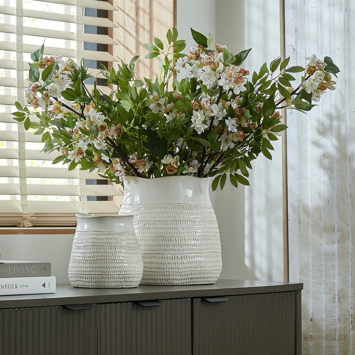 Hill Interiors Wide Hewn ceramic vases in white with textured rounded bodies, the larger filled with white blossom branches on a wooden sideboard by a window.