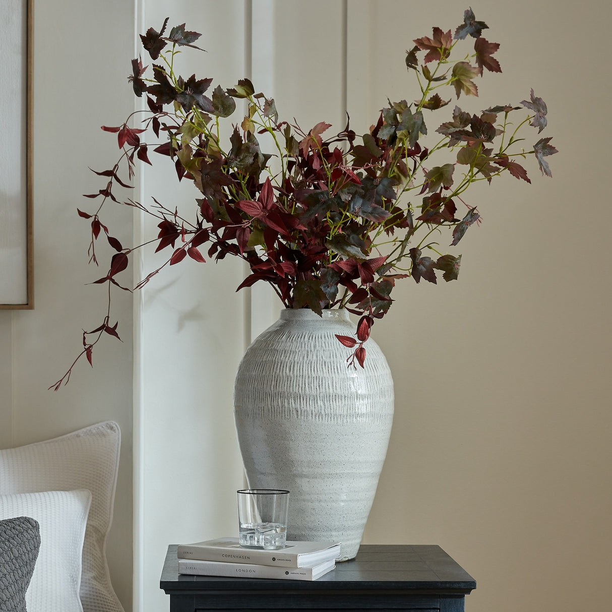 Hill Interiors Medium Olpe ceramic vase in white with textured ribbed body, holding tall burgundy foliage stems on a dark side table in a living room.