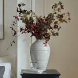 Hill Interiors Medium Olpe ceramic vase in white with textured ribbed body, holding tall burgundy foliage stems on a dark side table in a living room.