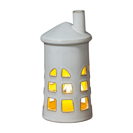 Hill Interiors Medium Lighthouse ceramic tealight holder in white with tapered tower, chimney top and cut-out windows glowing from a lit tealight inside.