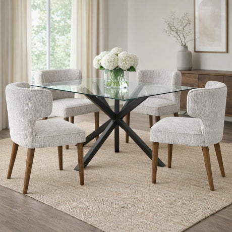 Dining room set with four Hill Interiors Quennomer grey upholstered wooden dining chairs around a square glass table on a black crossed base.