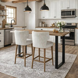 Hill Interiors Quennomer bar stools in grey textured upholstery with wooden legs, set at a wood and black metal island in a white farmhouse kitchen.