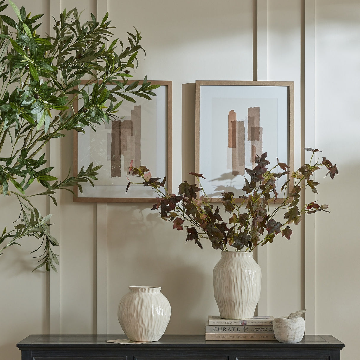 Hill Interiors framed wall art in wooden frames displayed above a dark sideboard with cream vases, foliage stems and stacked books in a neutral living space.