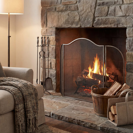 Hill Interiors large arched fire screen in antique silver metal, positioned against a stone fireplace with burning logs, tools and a wooden log basket.